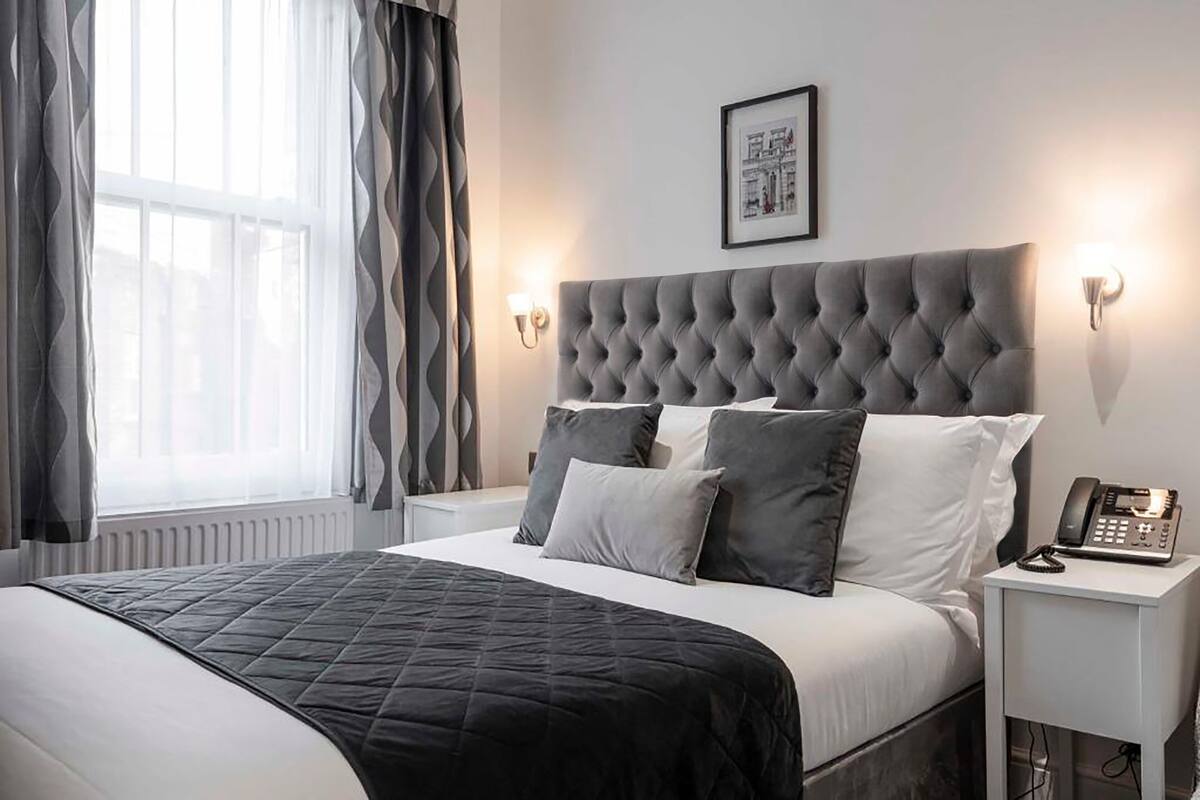 A neatly made bed with a grey headboard, white pillows, and a black bedspread. There is a bedside table with a telephone and a framed picture on the wall above it.