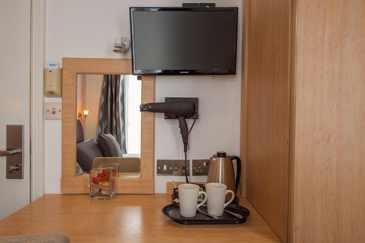 A hotel room with a television mounted on the wall, a hairdryer, a coffee maker, and a tray with two mugs.