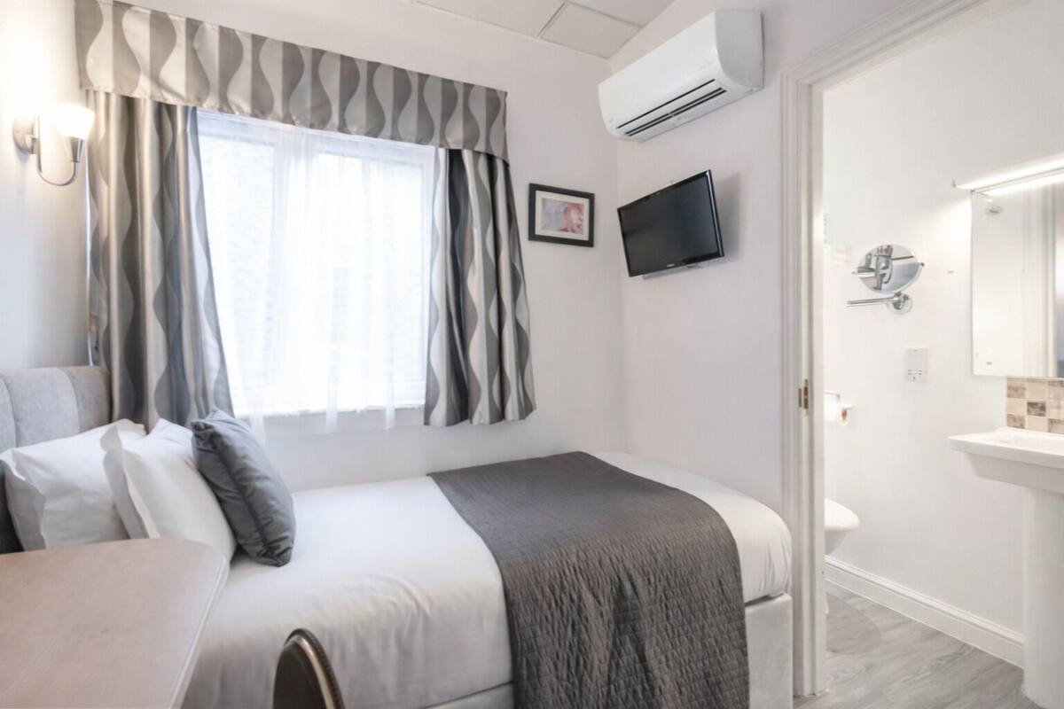 A hotel room with a bed, a TV, an air conditioner, a bathroom with a sink, and a window with curtains.