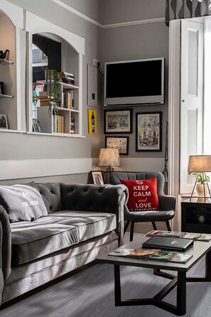 A modern living room with a grey sofa, a red cushion, a coffee table with magazines, a TV on the wall, and a bookshelf with various items. - The Melita (London)