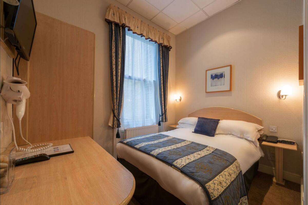 A hotel room with a bed, a desk with a telephone and remote control, a wall-mounted lamp, and a window with curtains.