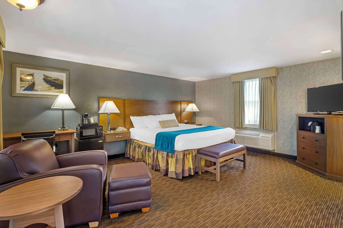 A hotel room with a bed, two bedside lamps, a TV, a desk with a lamp, a chair, a small table, and a bench.