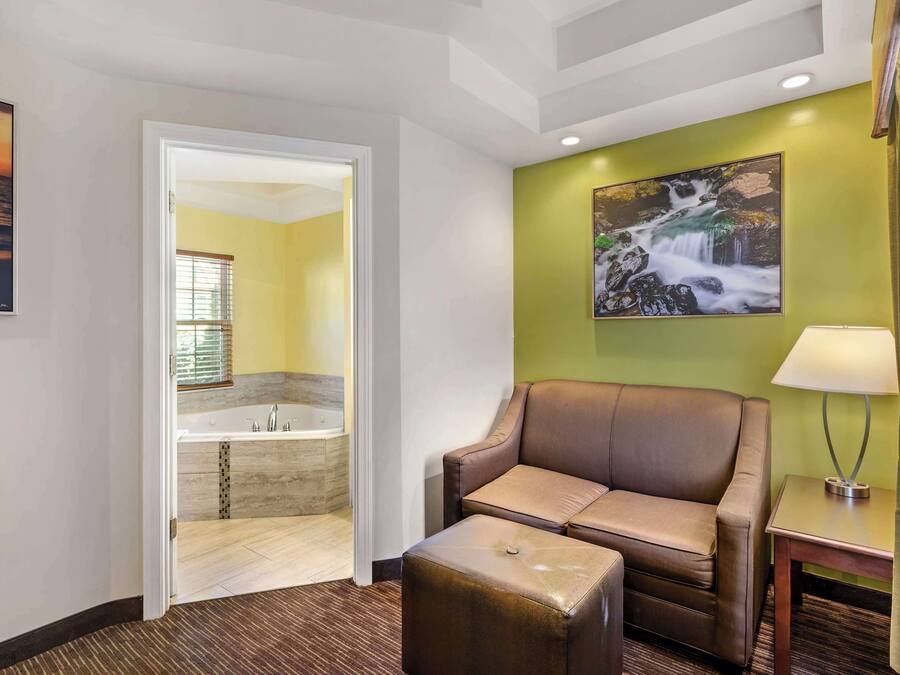 Une chambre d’hôtel avec un canapé en cuir marron, un petit pouf, une lampe fixée au mur et un tableau représentant une cascade.