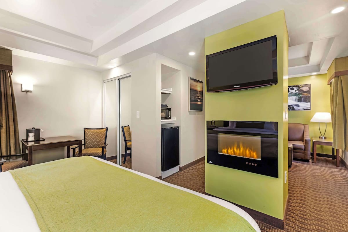 A hotel room with a bed, a flat-screen TV, a wall-mounted fireplace, a dining area with a table and chairs, and a small kitchen area.