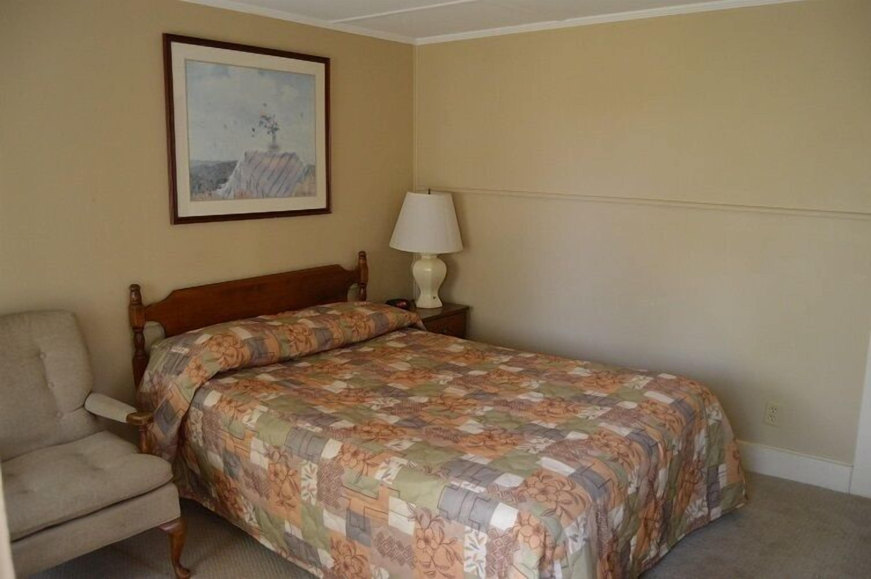 Un dormitorio con cama, una silla, una mesita de noche, una lámpara y un cuadro en la pared.