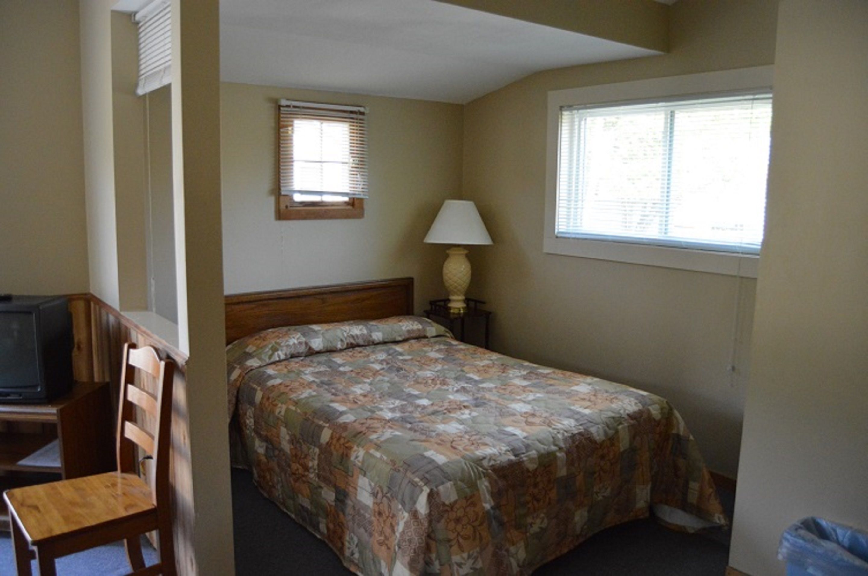 A bedroom with a bed, a chair, a desk with a television, a window with blinds, and a lamp.