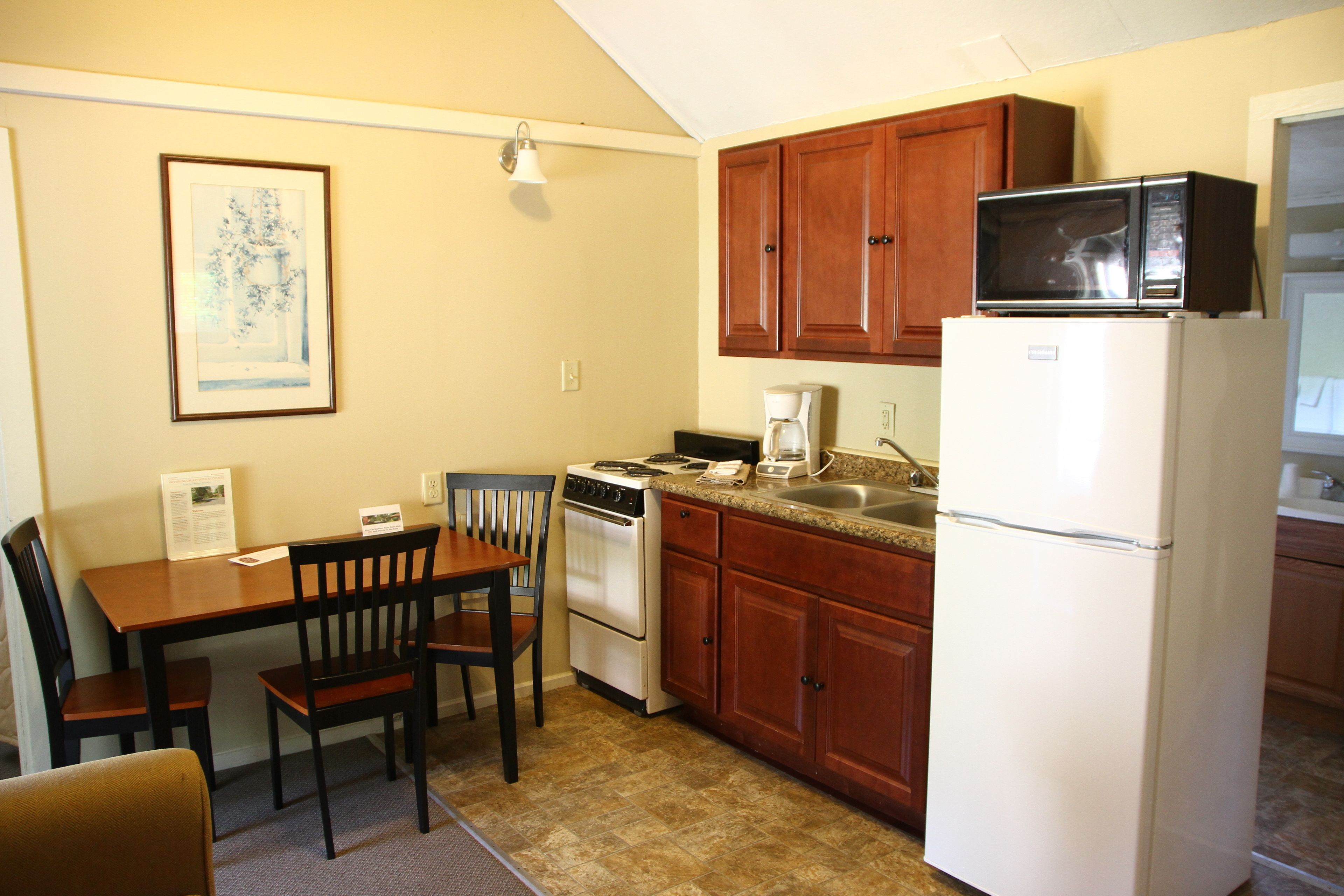 A compact kitchen with wooden cabinets, a white refrigerator, a microwave, a stove, and a dining table with chairs.
