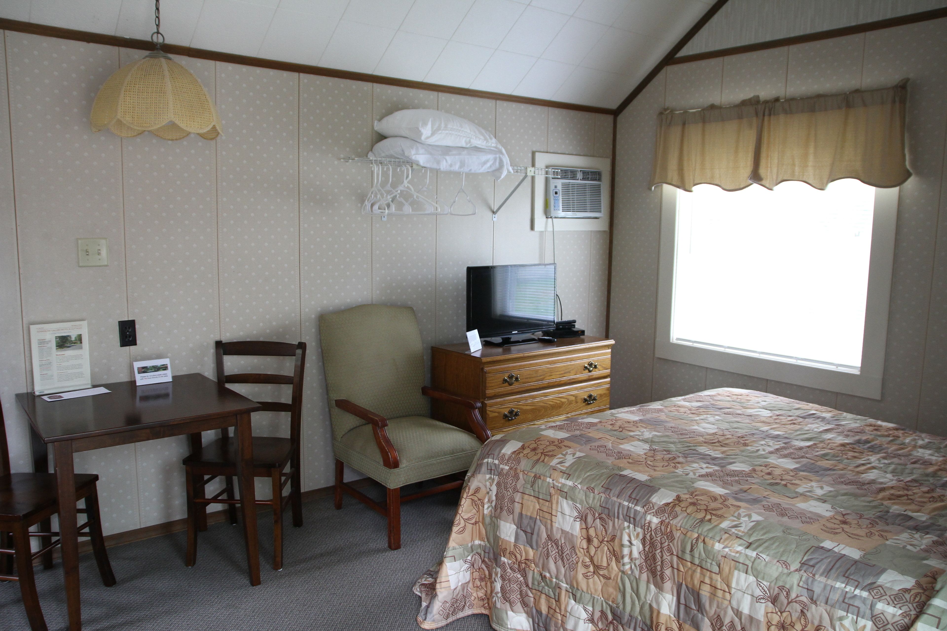 A hotel room with a bed, a television on a wooden dresser, a chair, a desk, and a window with curtains.