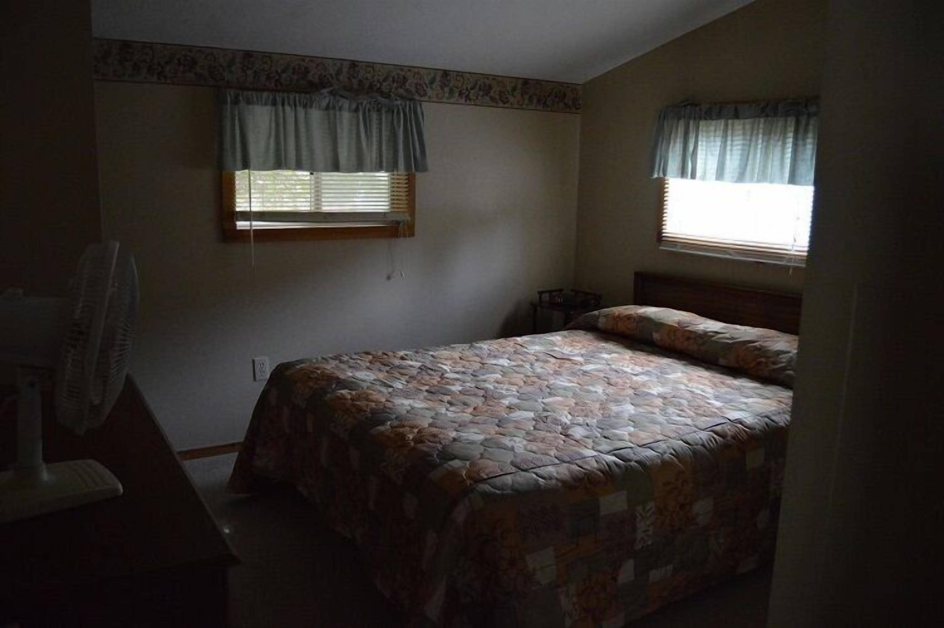 A bedroom with a bed, two windows with curtains, a fan, and a dresser.