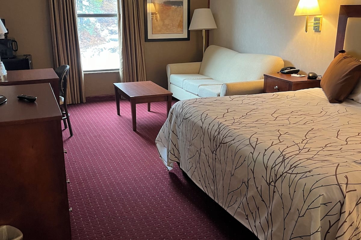 A hotel room with a large bed, a desk, a chair, a small table, and a window with curtains.