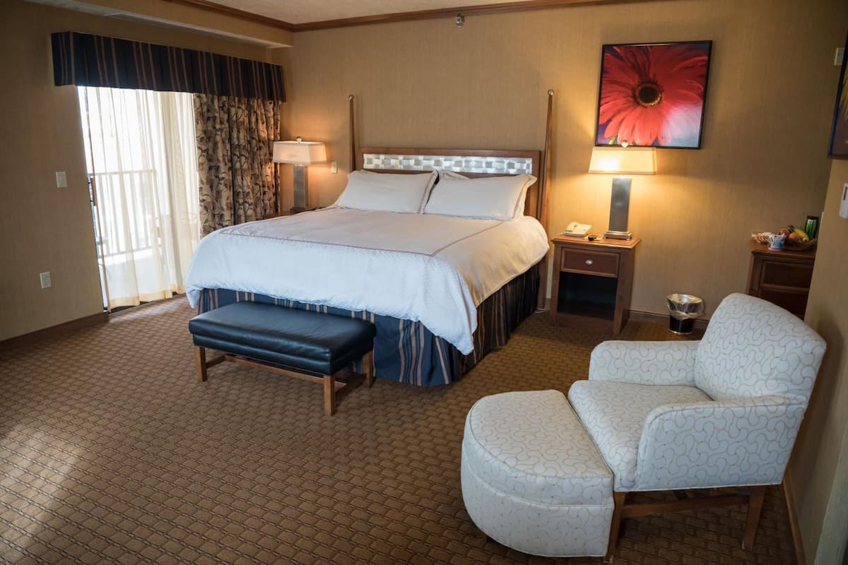A hotel room with a large bed, a bench, a chair, a bedside table, a lamp, and a painting on the wall.