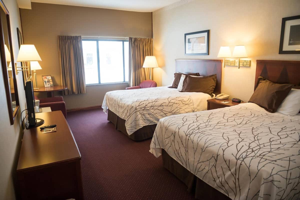 A hotel room with two beds, a desk, a chair, a window with curtains, and two wall-mounted lamps.
