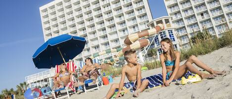 A beach scene with a large hotel in the background, a blue umbrella, and beach chairs.