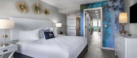 A modern hotel room with a large bed, bedside lamps, a desk, and a view of the ocean through a glass door.