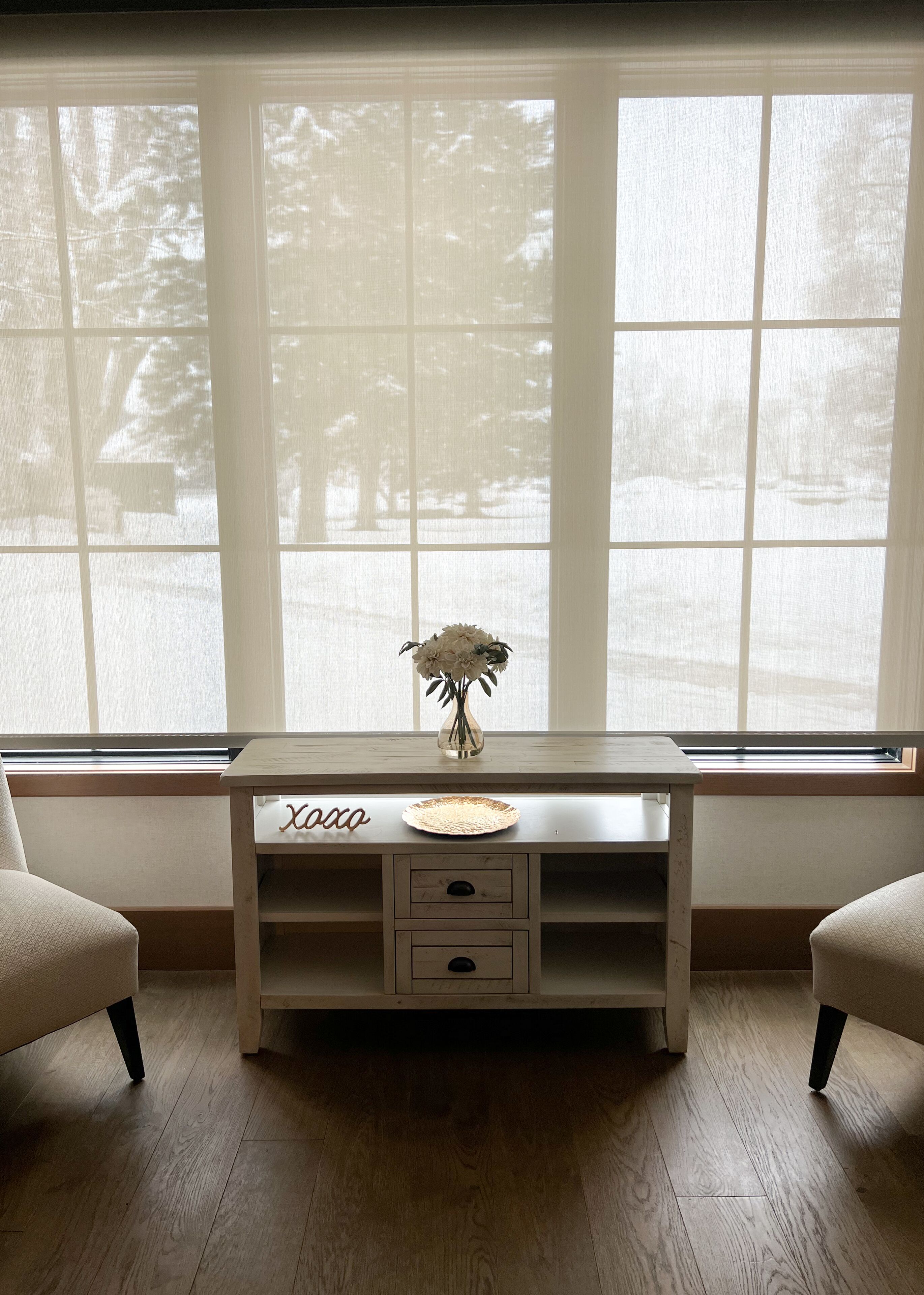 A wooden console table with a vase of flowers, a plate, and a sign reading "xoxo" in a room with large windows and a view of trees.