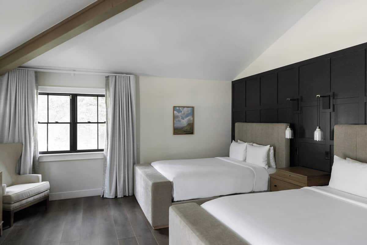 Ein Hotelzimmer mit zwei Betten, einem Sessel, einem Fenster mit Vorhängen, einem Gemälde und einem dunklen Kopfteil.