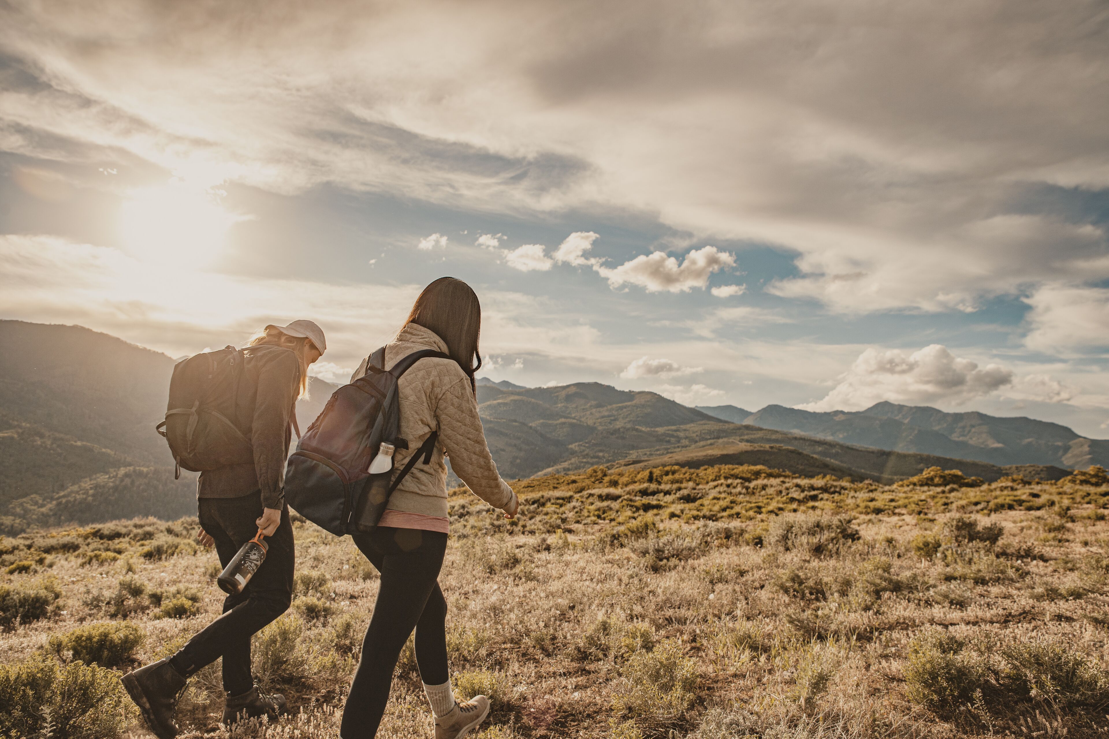 Two people hiking on a grassy field with mountains in the background.