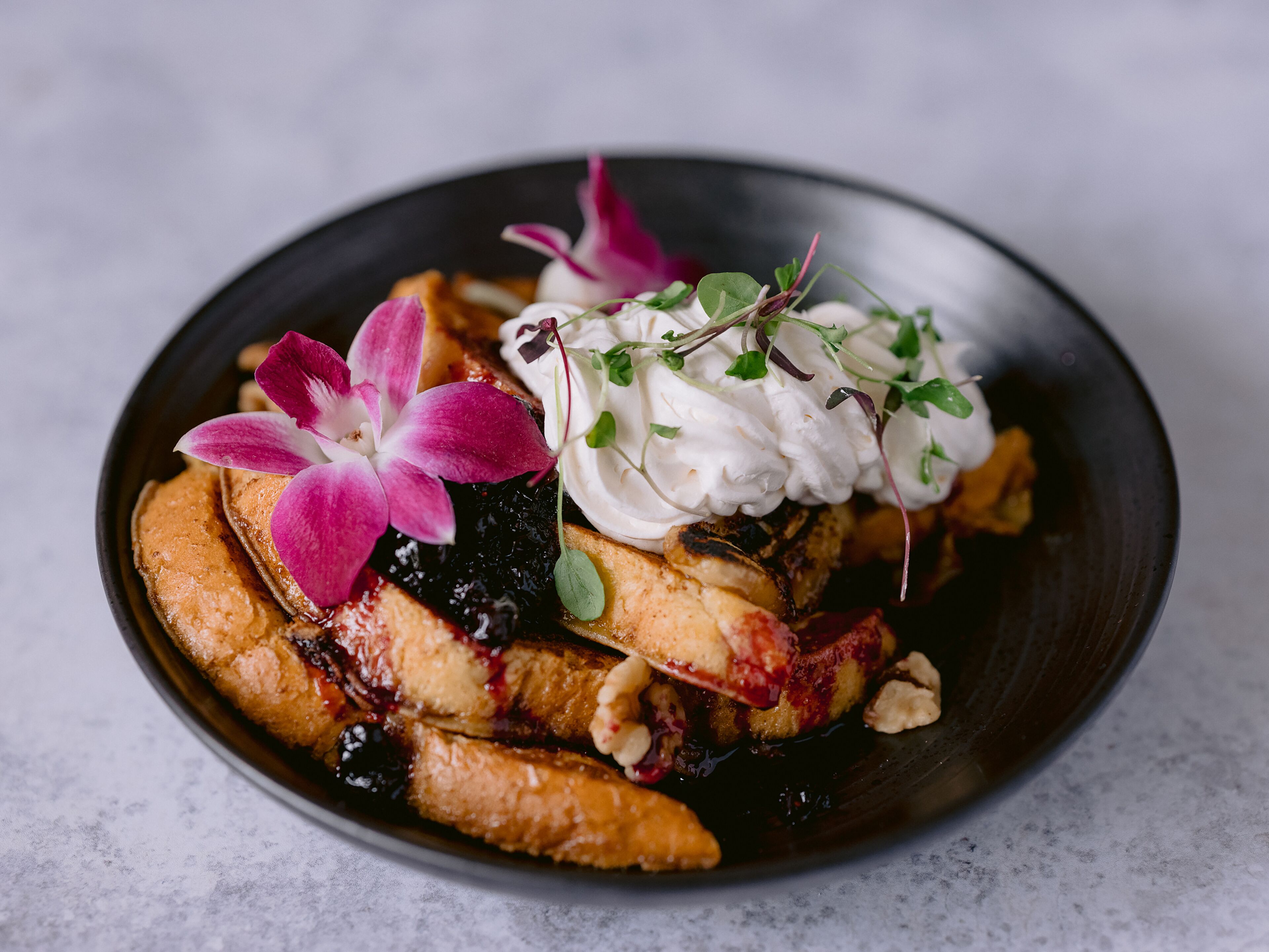 Un plato de postre con panqueques, frutos rojos, crema batida y una orquídea rosa.