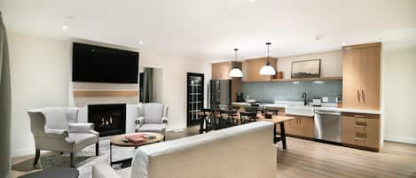 A modern living room with a sofa, armchairs, and a coffee table. A fireplace is visible in the background. The kitchen features wooden cabinets, a sink, and a dishwasher.
