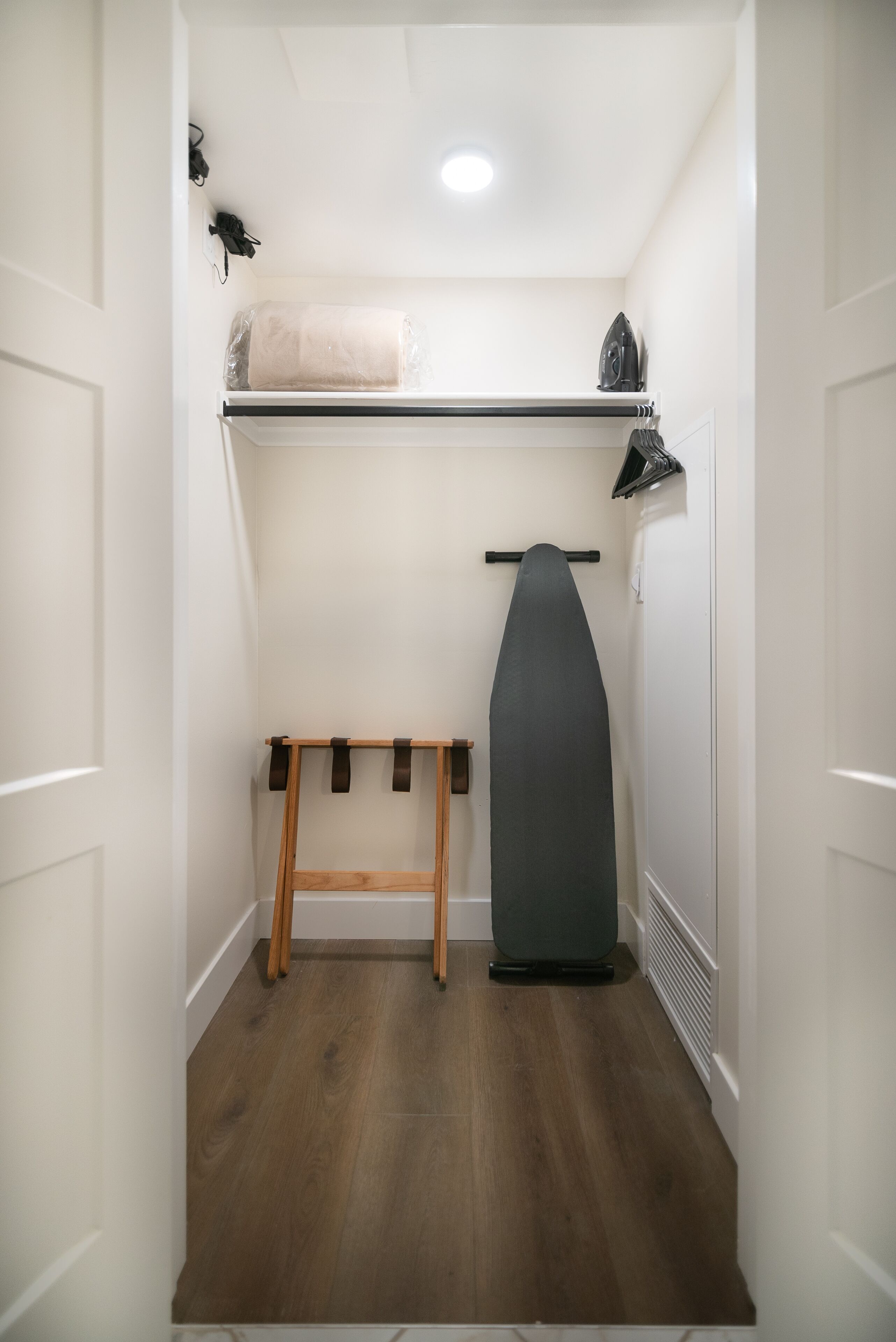 A small, narrow room with a wooden floor, a black ironing board, and a large, dark-colored iron on the wall.