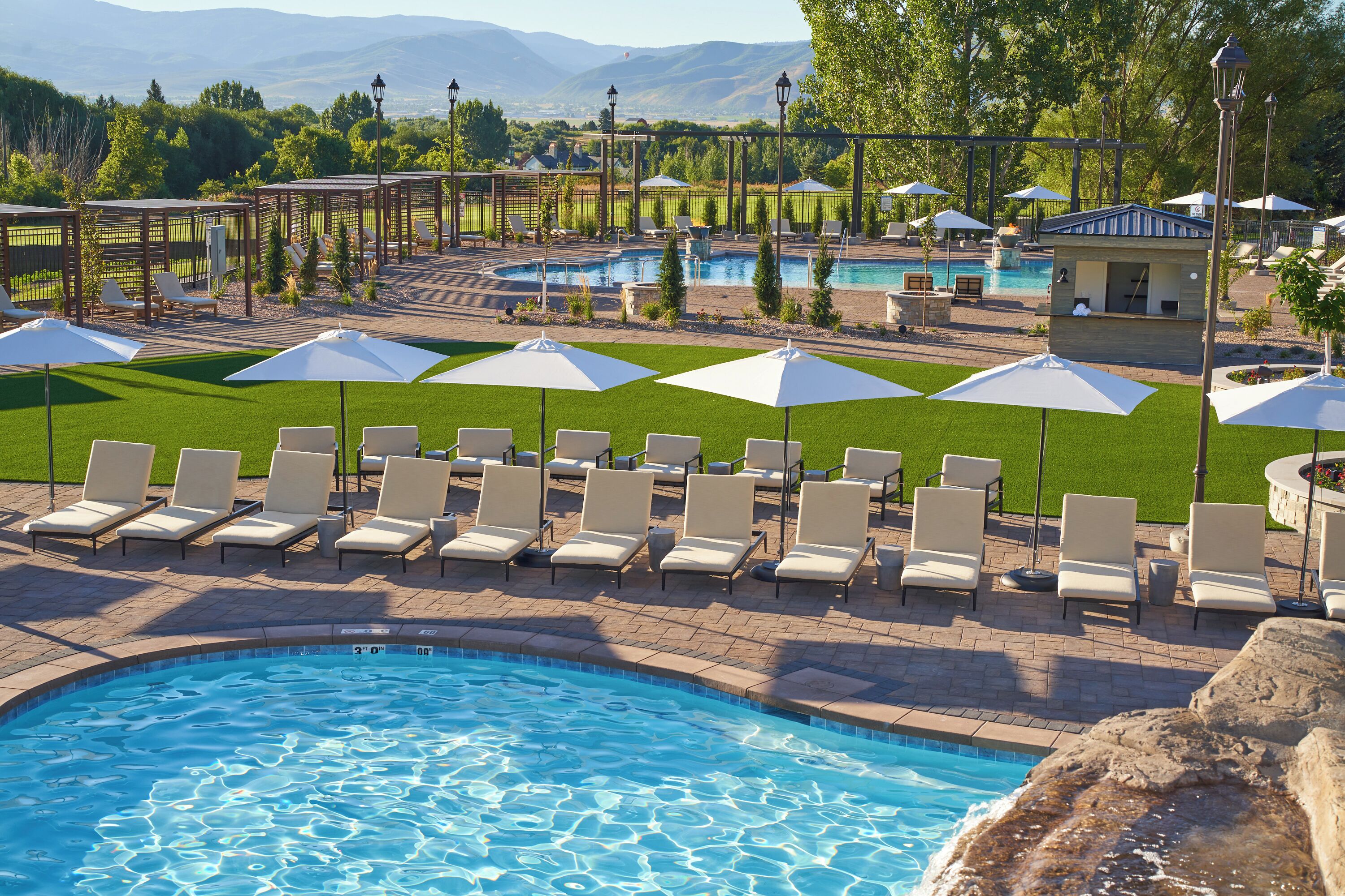 A pool area with lounge chairs and umbrellas, surrounded by a landscaped garden and a view of mountains.