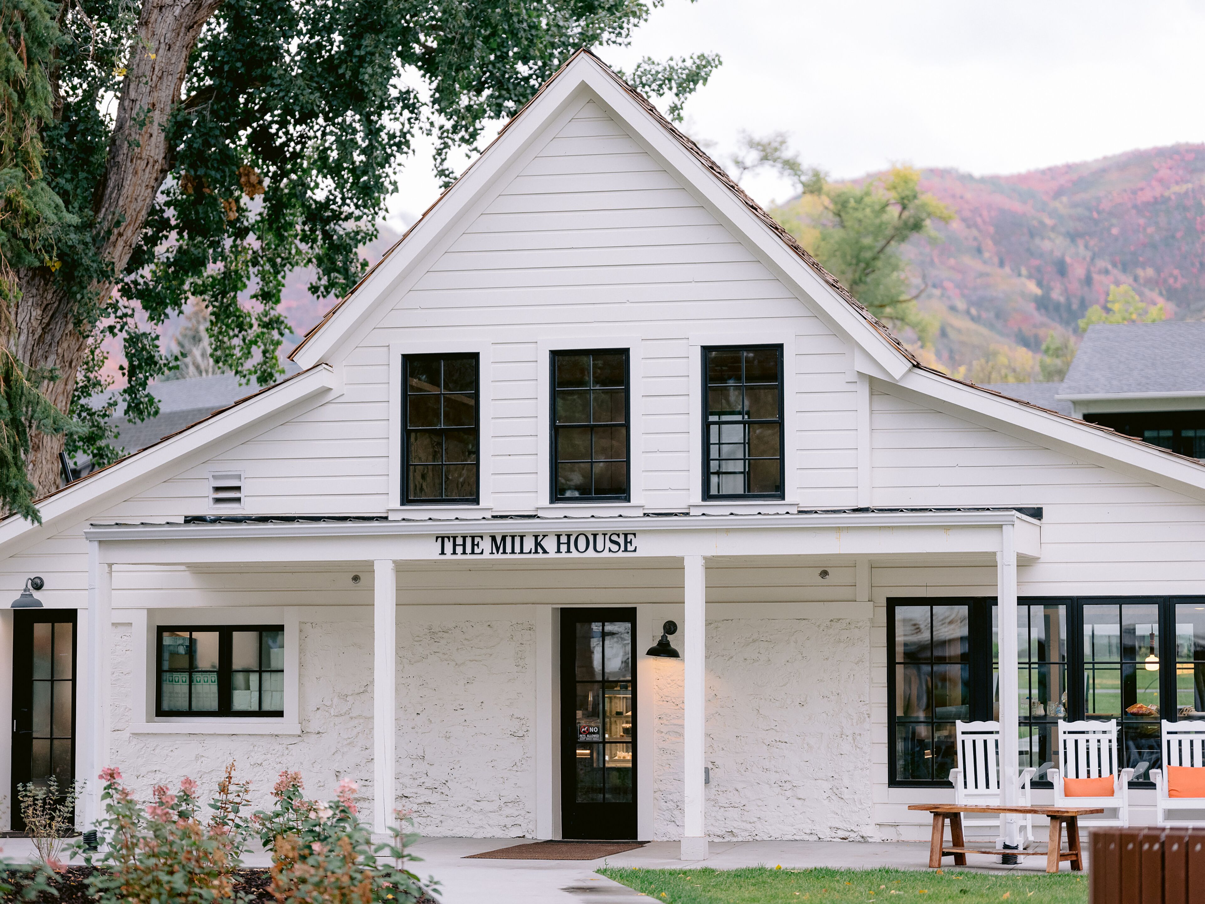 A white building with a sign reading "The Milk House." It features a front porch with white chairs and a table.