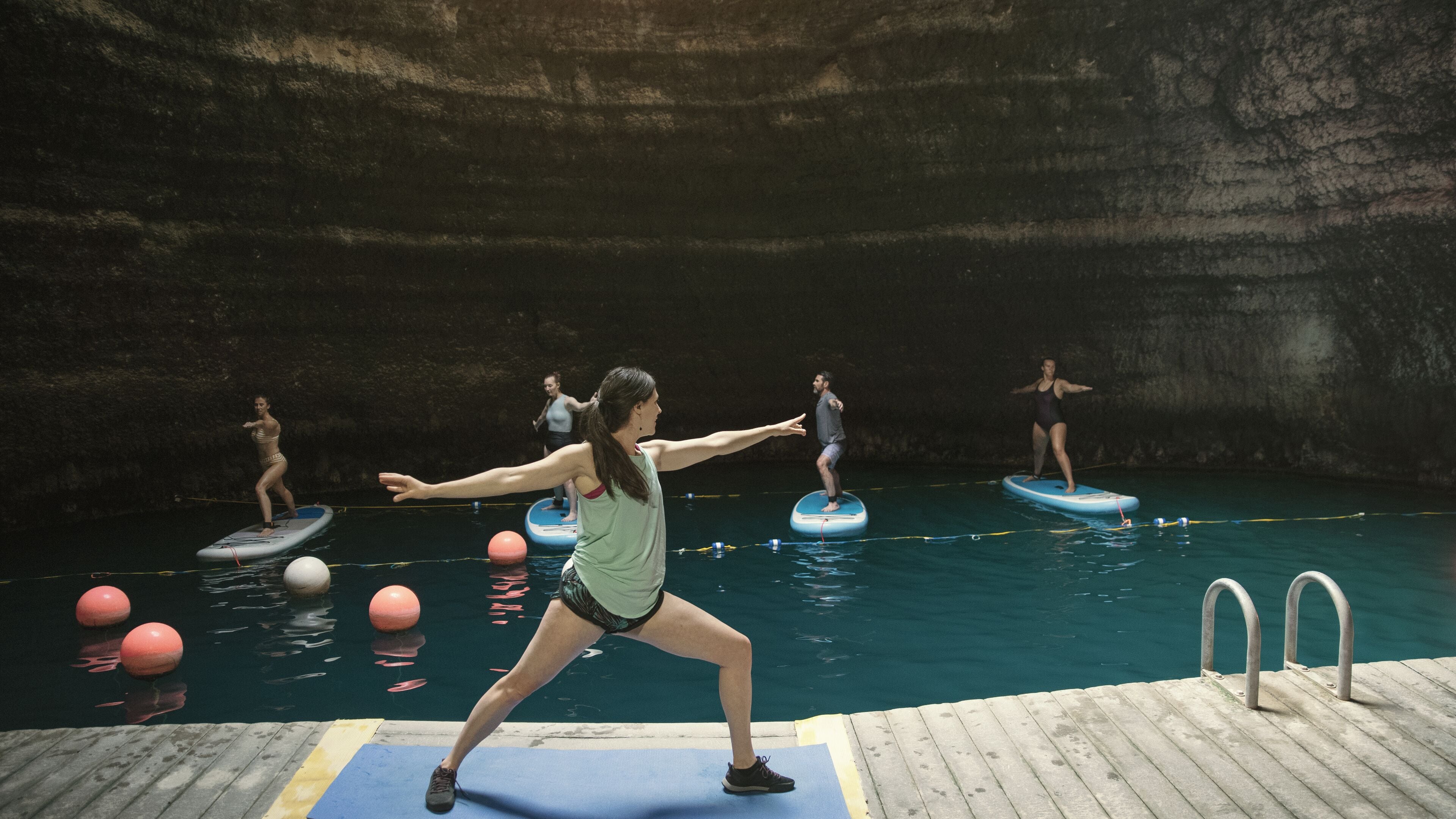 A group of people practicing on balance boards in a pool with a rocky wall in the background.
