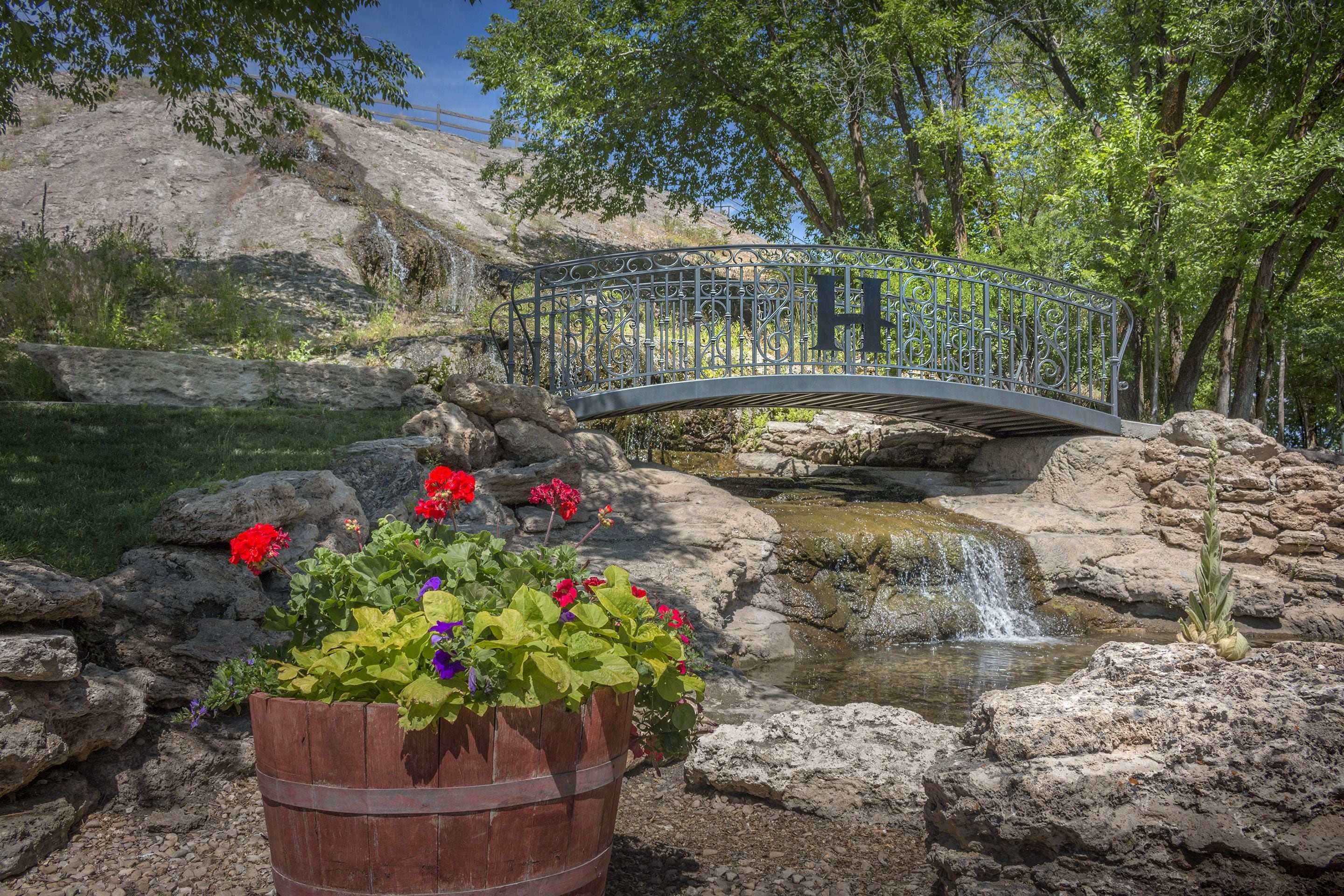 Una jardinera de madera con flores, una cascada pequeña y un puente metálico sobre un arroyo en un entorno natural.