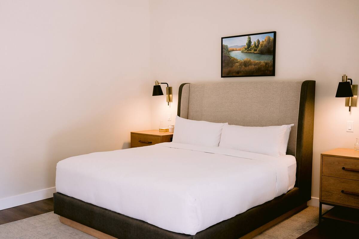 Ein ordentlich bezogenes Bett mit Kopfteil, ein Nachttisch mit Lampe und ein an der Wand hängendes Bild.