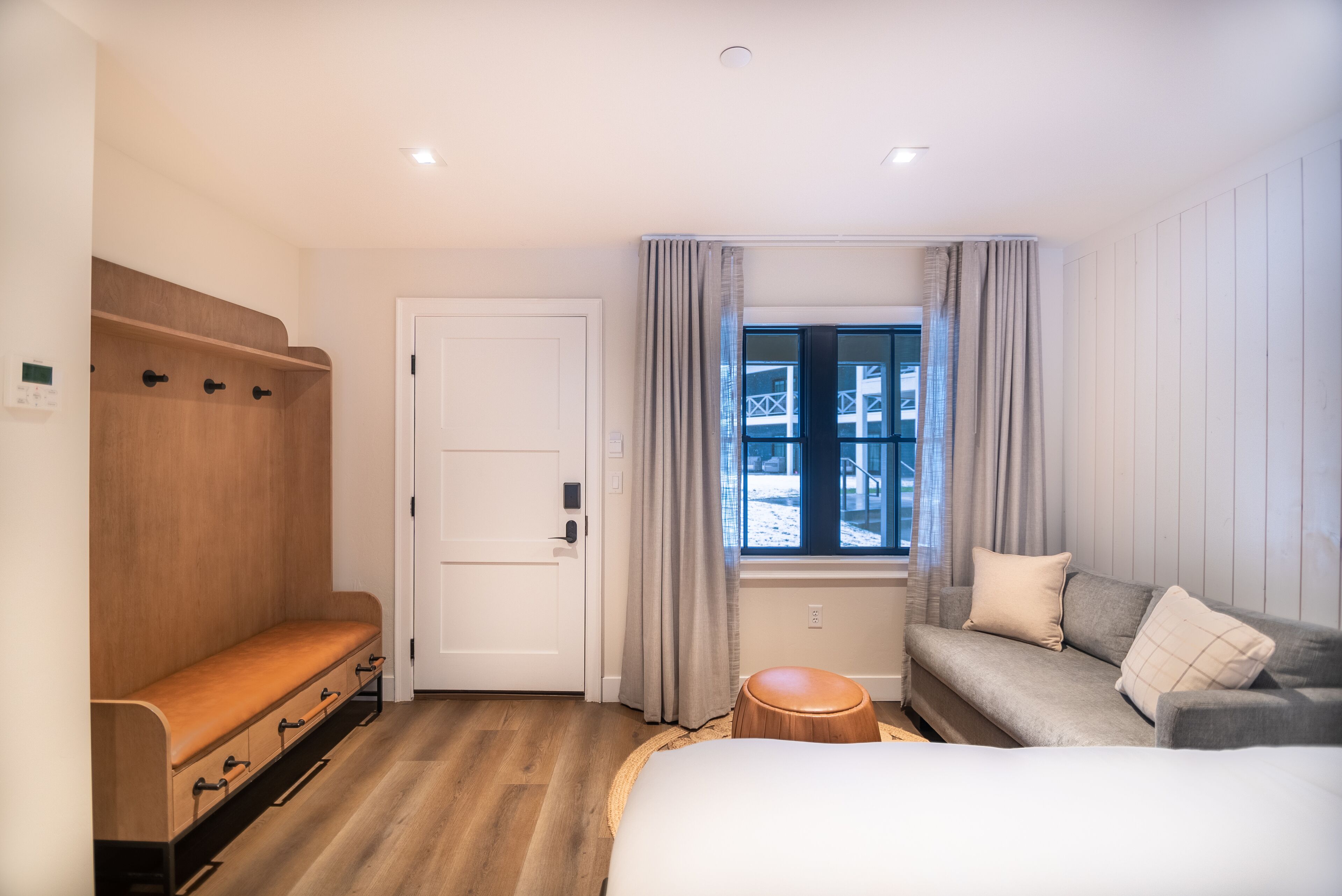 A hotel room with a wooden bench, a sofa, a bed, a window with curtains, and a door.