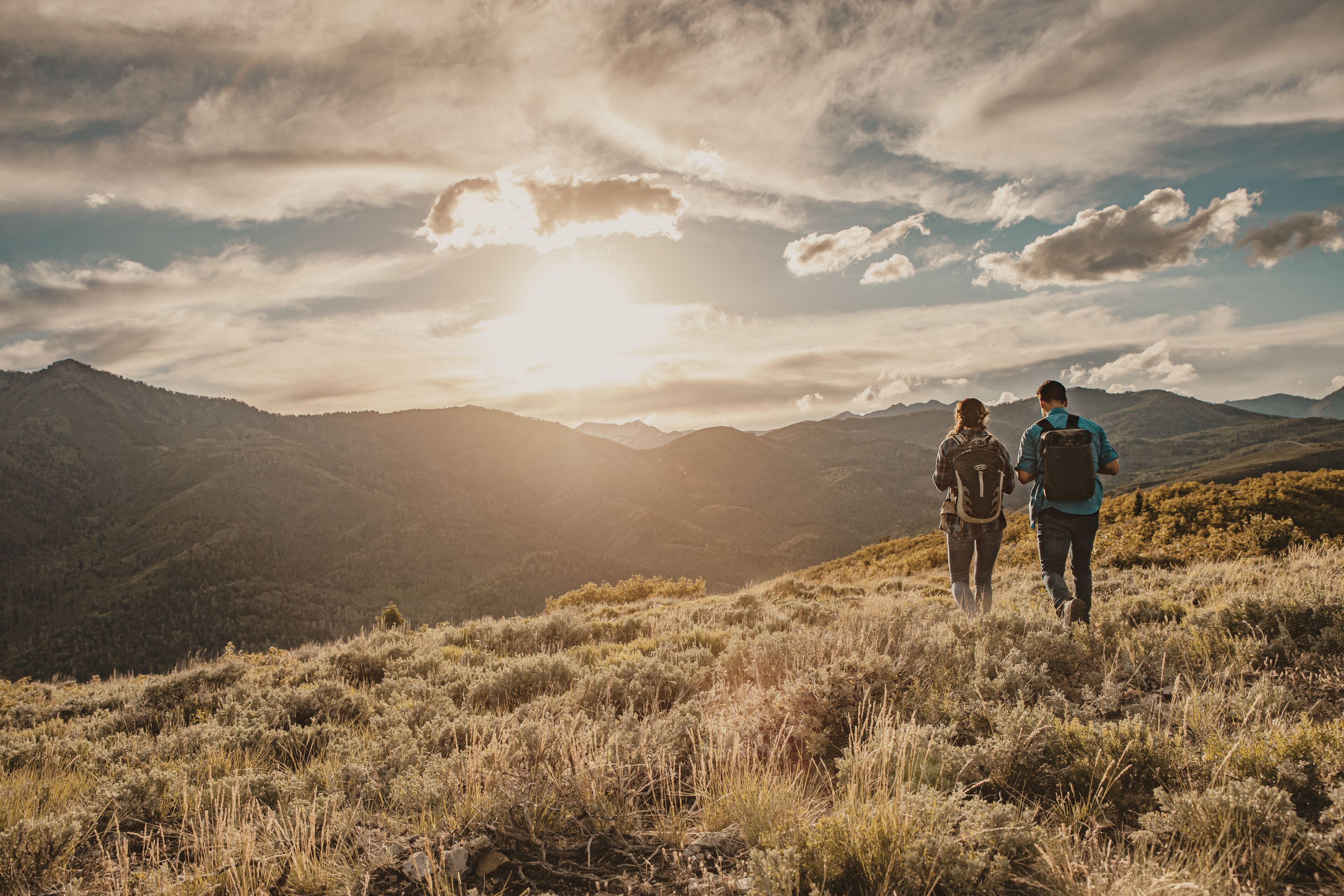 Two people walking on a grassy hillside with mountains in the background and the sun setting behind them.