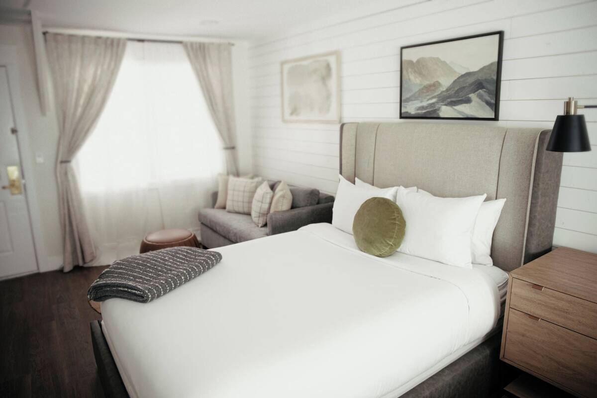 Ein Hotelzimmer mit Bett, Sofa, Nachttisch, Lampe und einem Fenster mit Vorhängen.