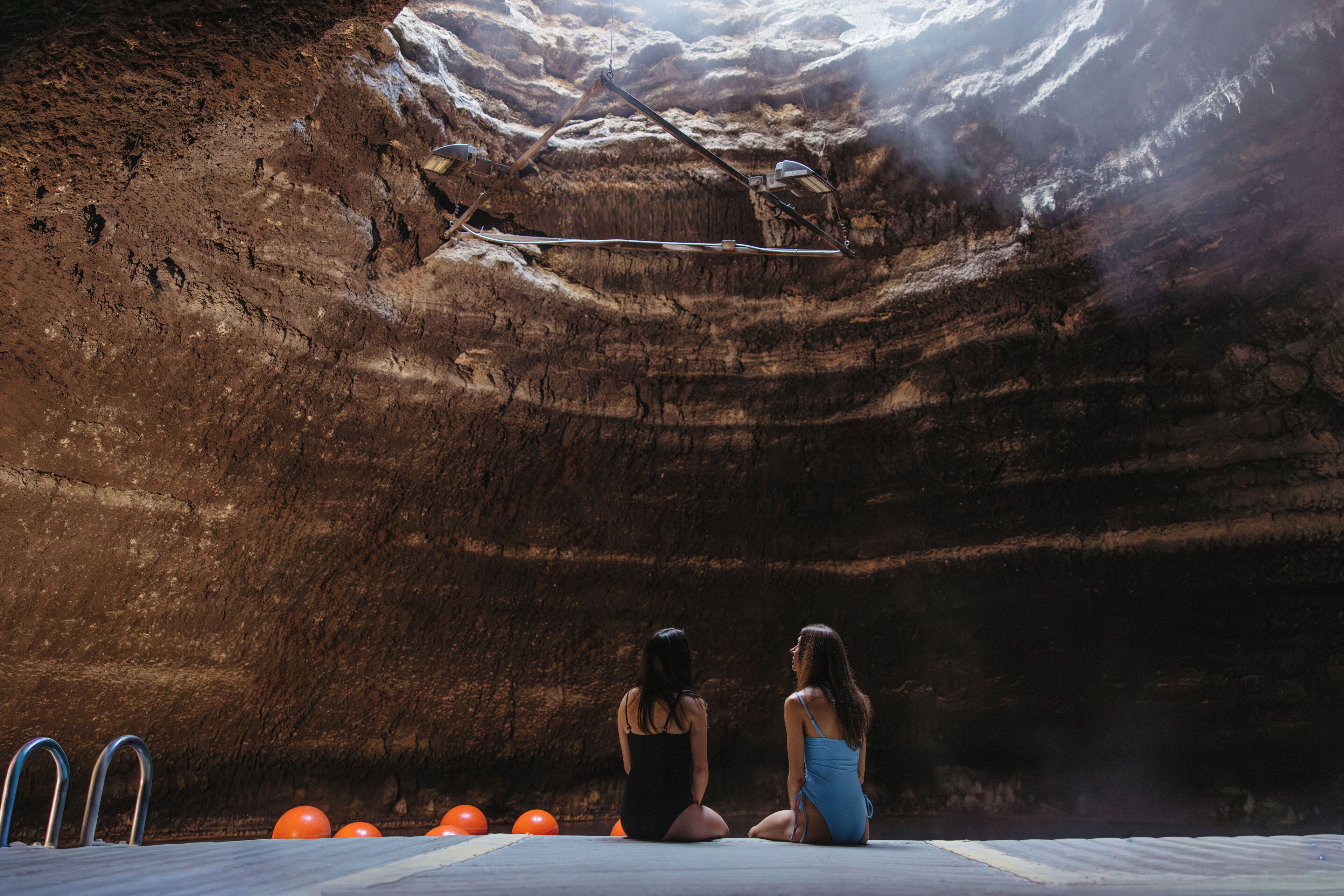 Two women sitting on a ledge in a canyon with a metal structure above them.