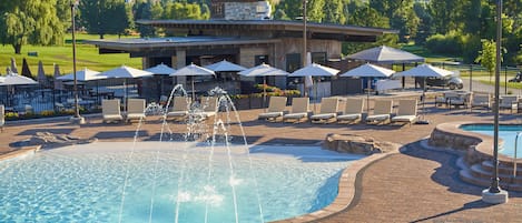 A pool area with a water fountain, lounge chairs, umbrellas, and a building with a stone facade in the background.