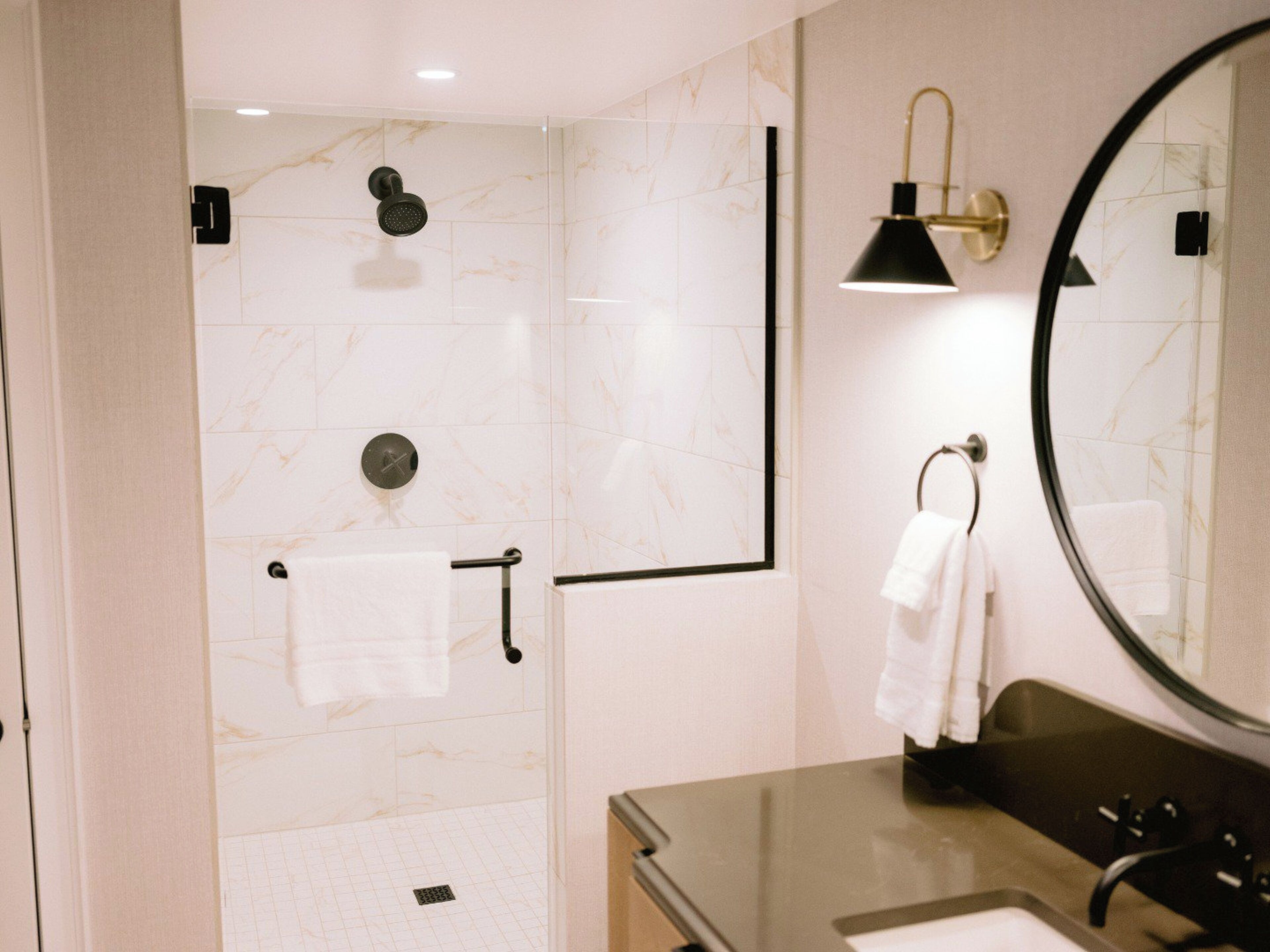 A modern bathroom with a glass shower enclosure, a round mirror, a sink, and a towel rack.