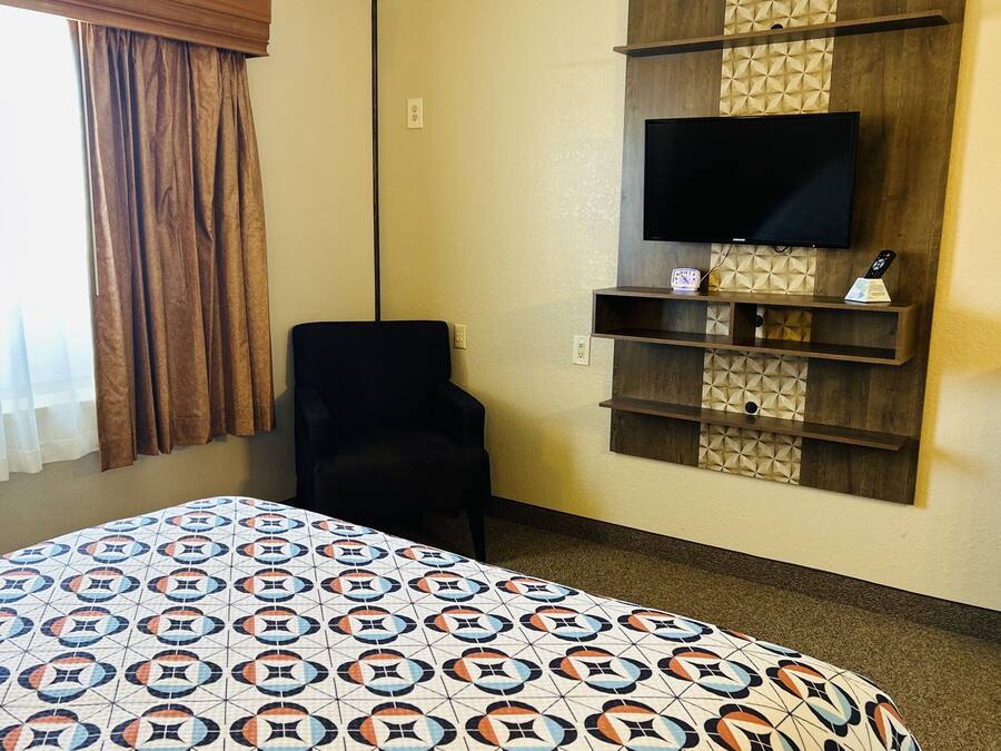 A hotel room with a large bed, a TV mounted on a wooden panel, a chair, and a small shelf with a clock and remote.