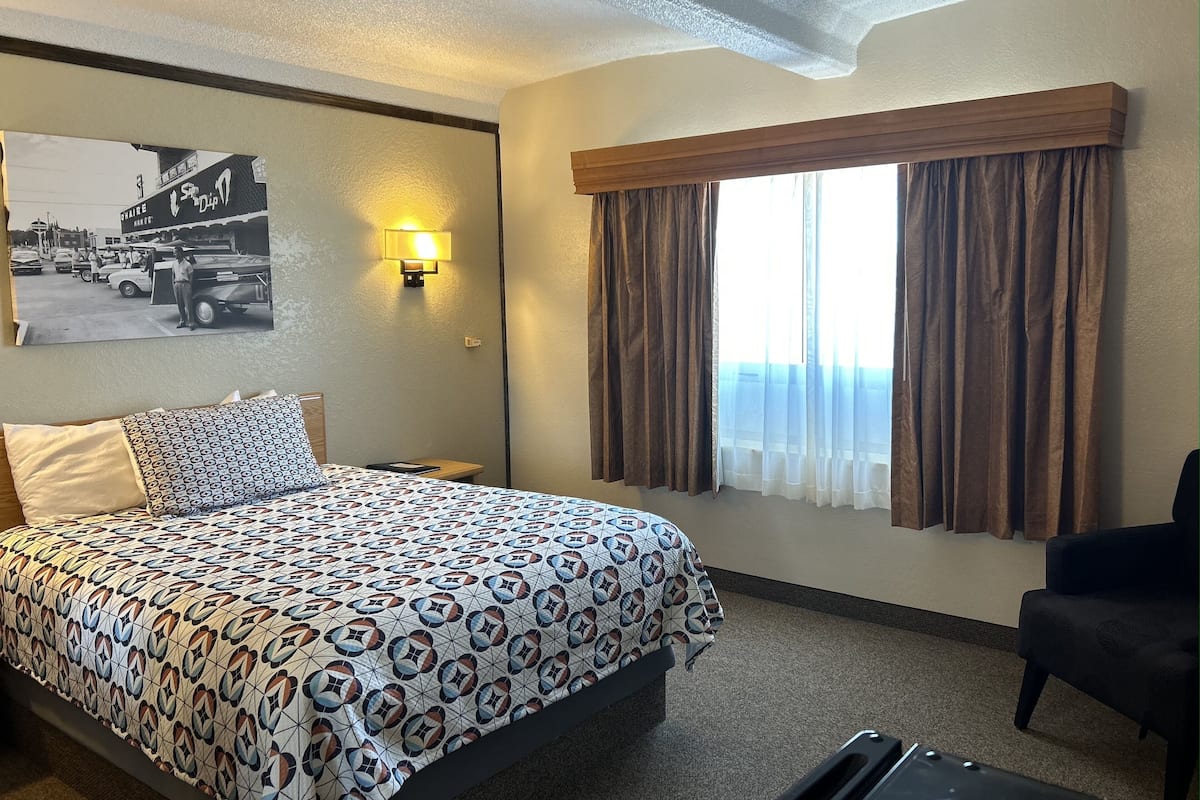 A hotel room with a bed, a nightstand, a lamp, a window with curtains, and a wall-mounted picture.
