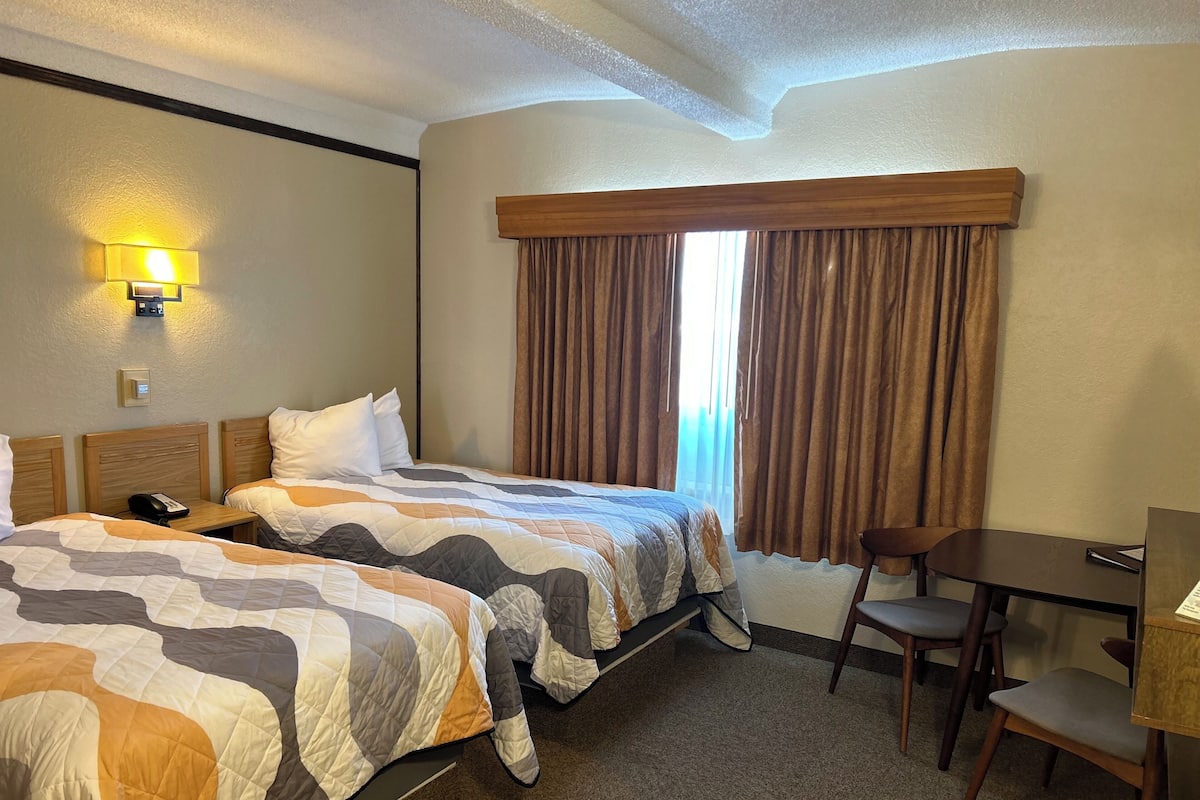 A hotel room with two beds, a desk, a chair, a lamp, and a window with curtains.