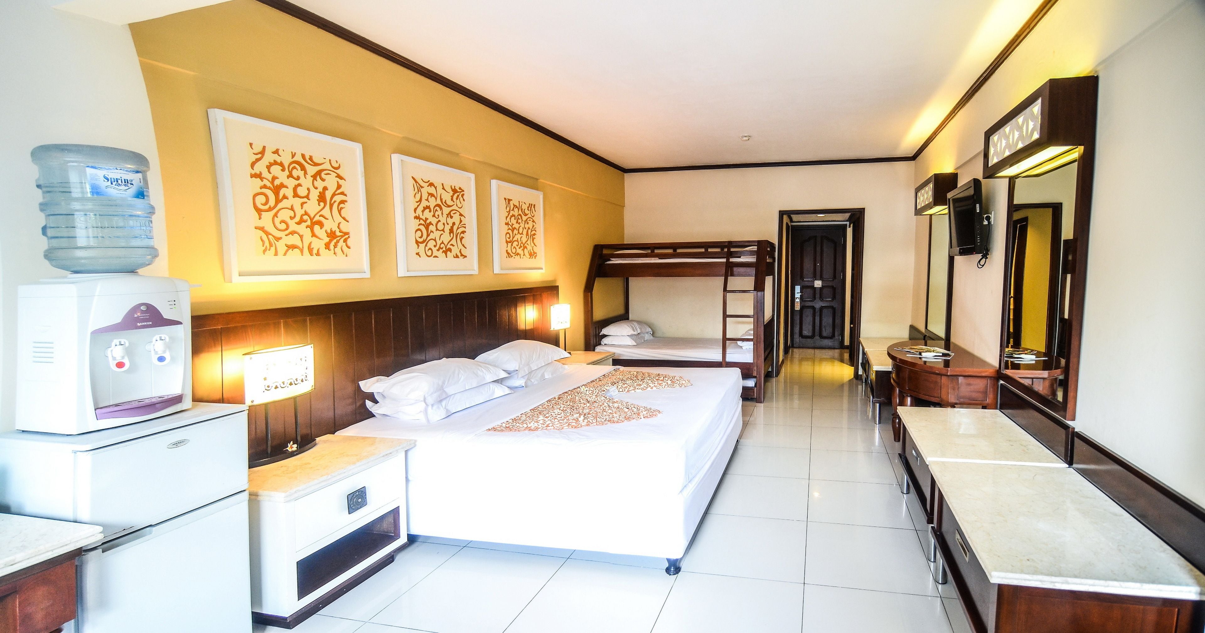 A hotel room with a bed, bedside tables, a water dispenser, a refrigerator, a mirror, and decorative wall art.