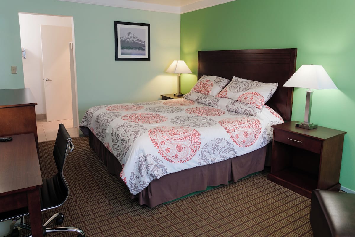 A hotel room with a bed, two bedside tables with lamps, a desk, a chair, and a door.