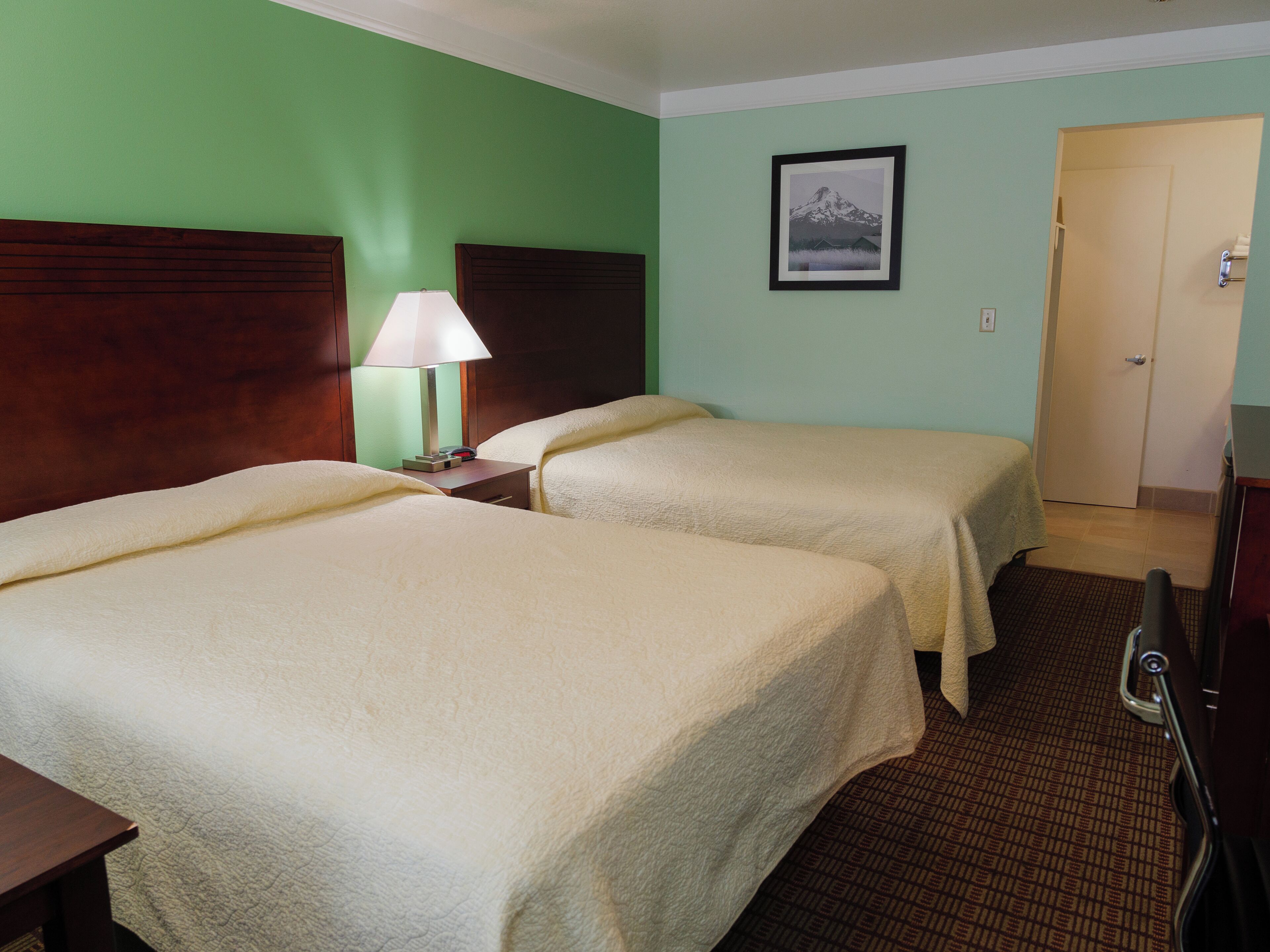 A hotel room with two beds, a nightstand with a lamp, a framed picture on the wall, and a door to the right.