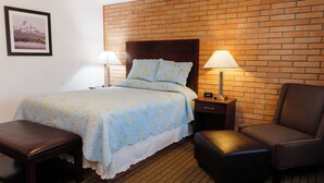A hotel room with a brick accent wall, a bed with floral bedding, two bedside lamps, a wooden nightstand, a brown leather armchair, and a small wooden bench. - Sweet Home Inn (Sweet Home)