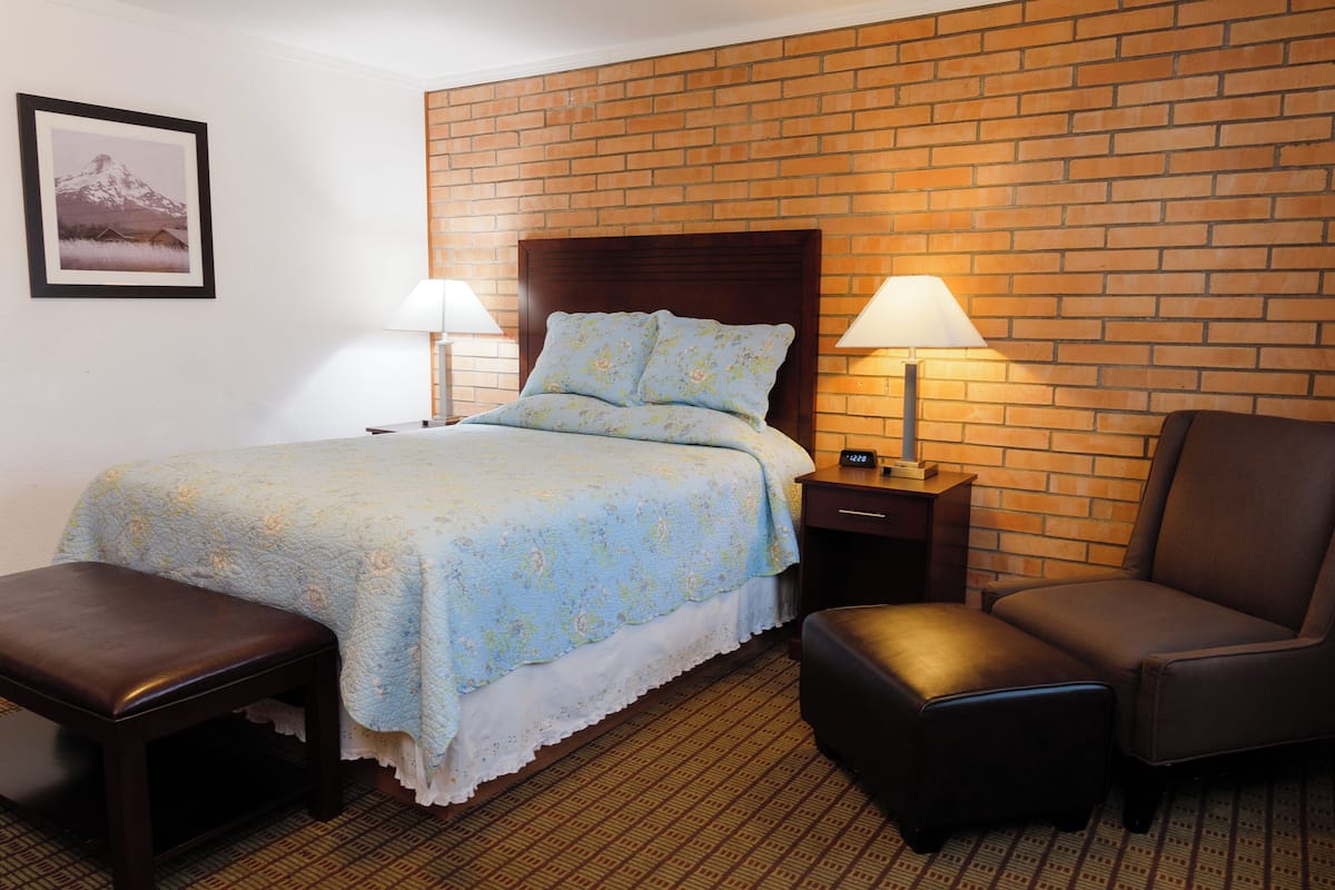 A hotel room with a brick accent wall, a bed with floral bedding, two bedside lamps, a wooden nightstand, a brown leather armchair, and a small wooden bench.