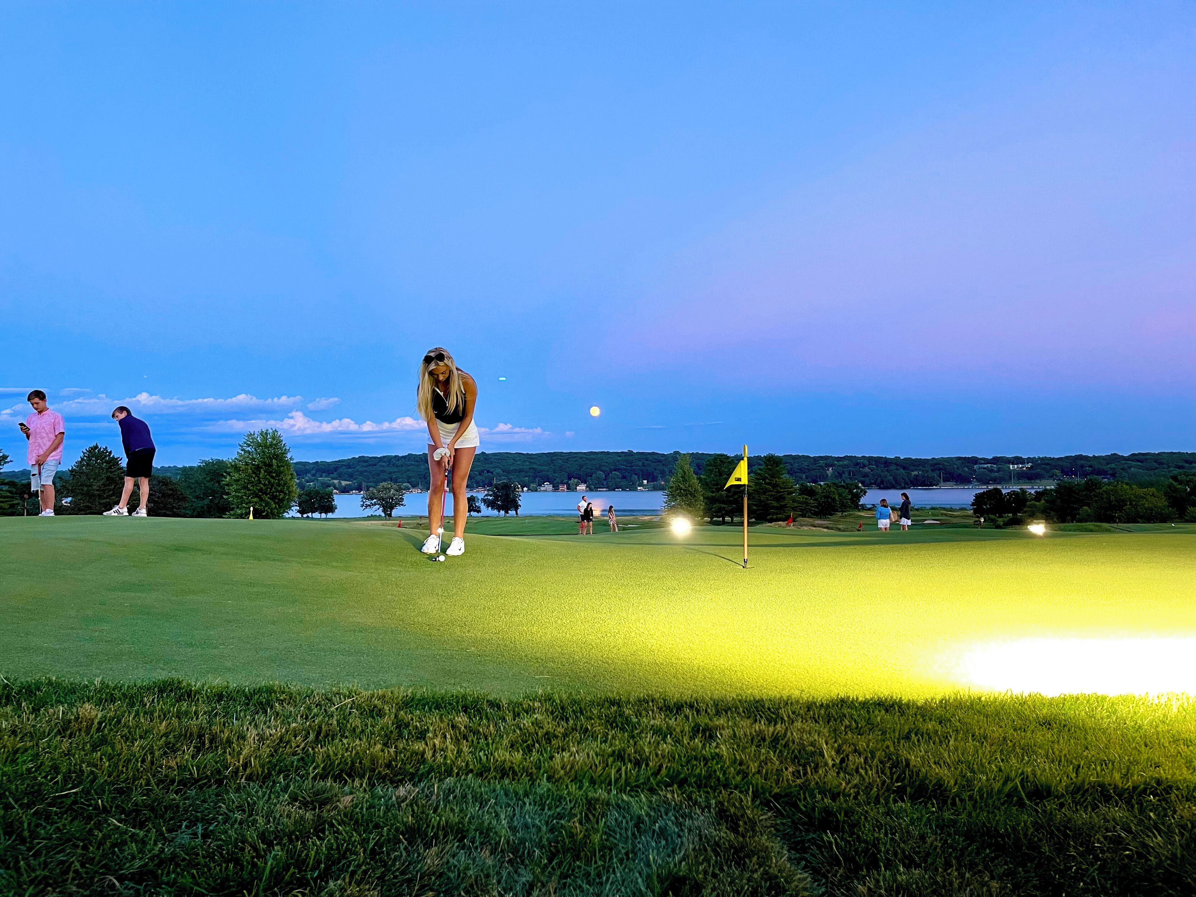 A golf course at dusk with players on the green and a body of water in the background.