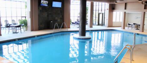 An indoor swimming pool with clear blue water, surrounded by seating areas and a large tree in the center.