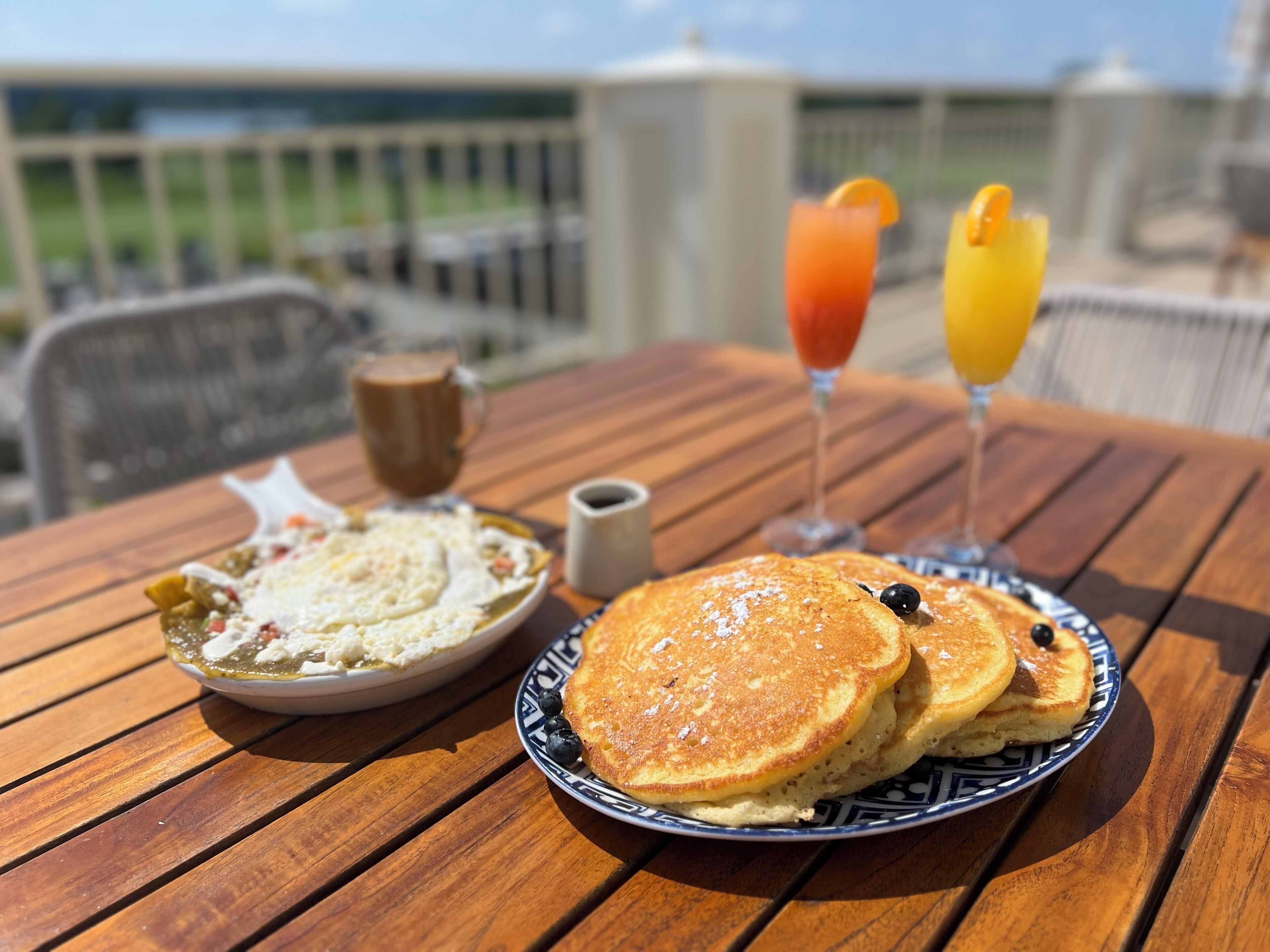 Una mesa de desayuno con panqueques, un plato de cereal, una taza de café y dos bebidas sobre una mesa de madera al aire libre.