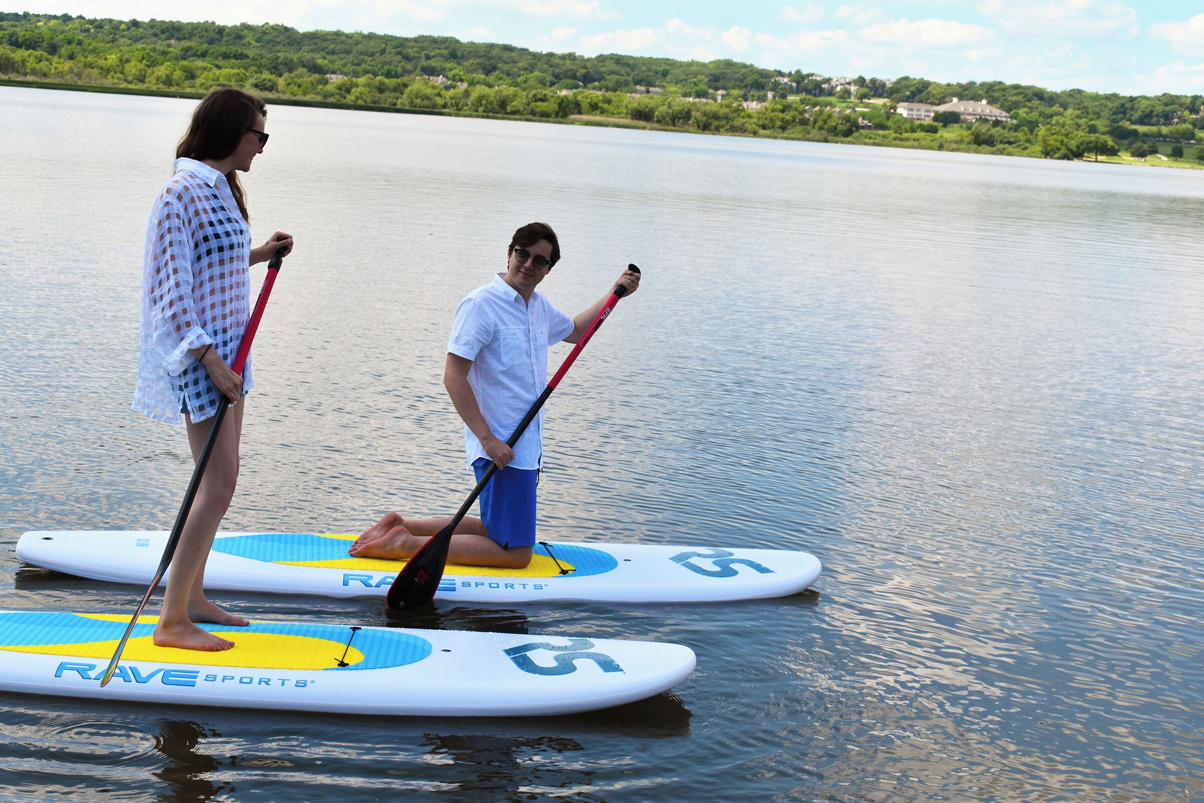 Twee mensen staan op paddleboards op een meer, met huizen op de achtergrond.