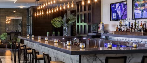 A modern bar area with a marble countertop, multiple bar stools, hanging light fixtures, and a television screen displaying a sports event.