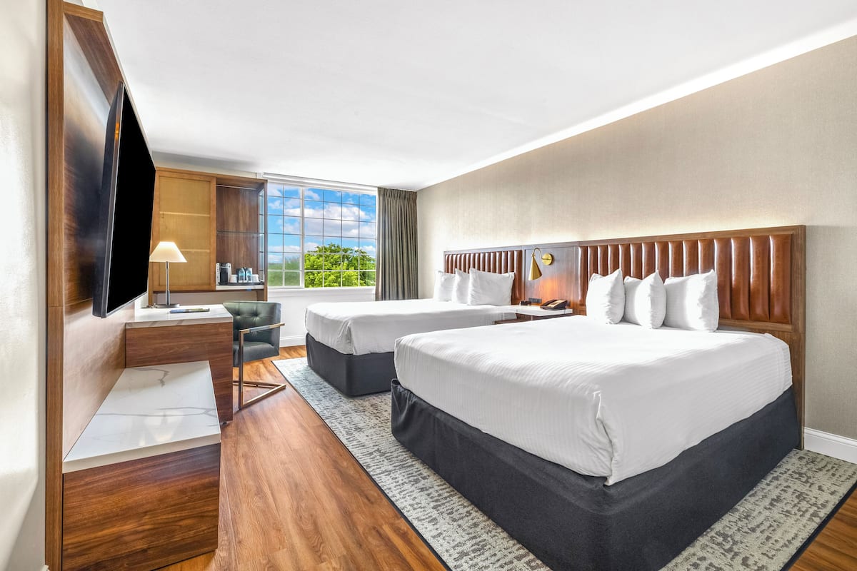 A hotel room with two beds, a desk, a TV, and a large window with a view of greenery.