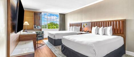 Une chambre d’hôtel avec deux lits, un bureau, une télévision et une grande fenêtre donnant sur un paysage verdoyant.